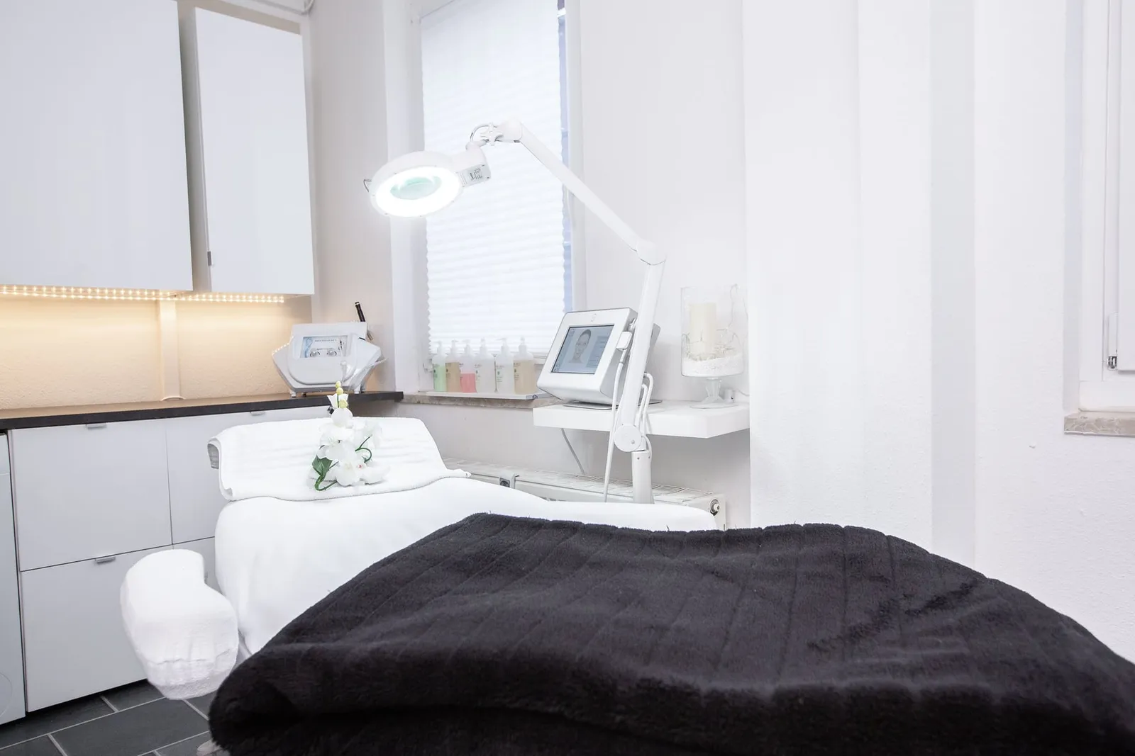 Behandlungsliege mit dunkler Decke, Lupenleuchte und Gerät.