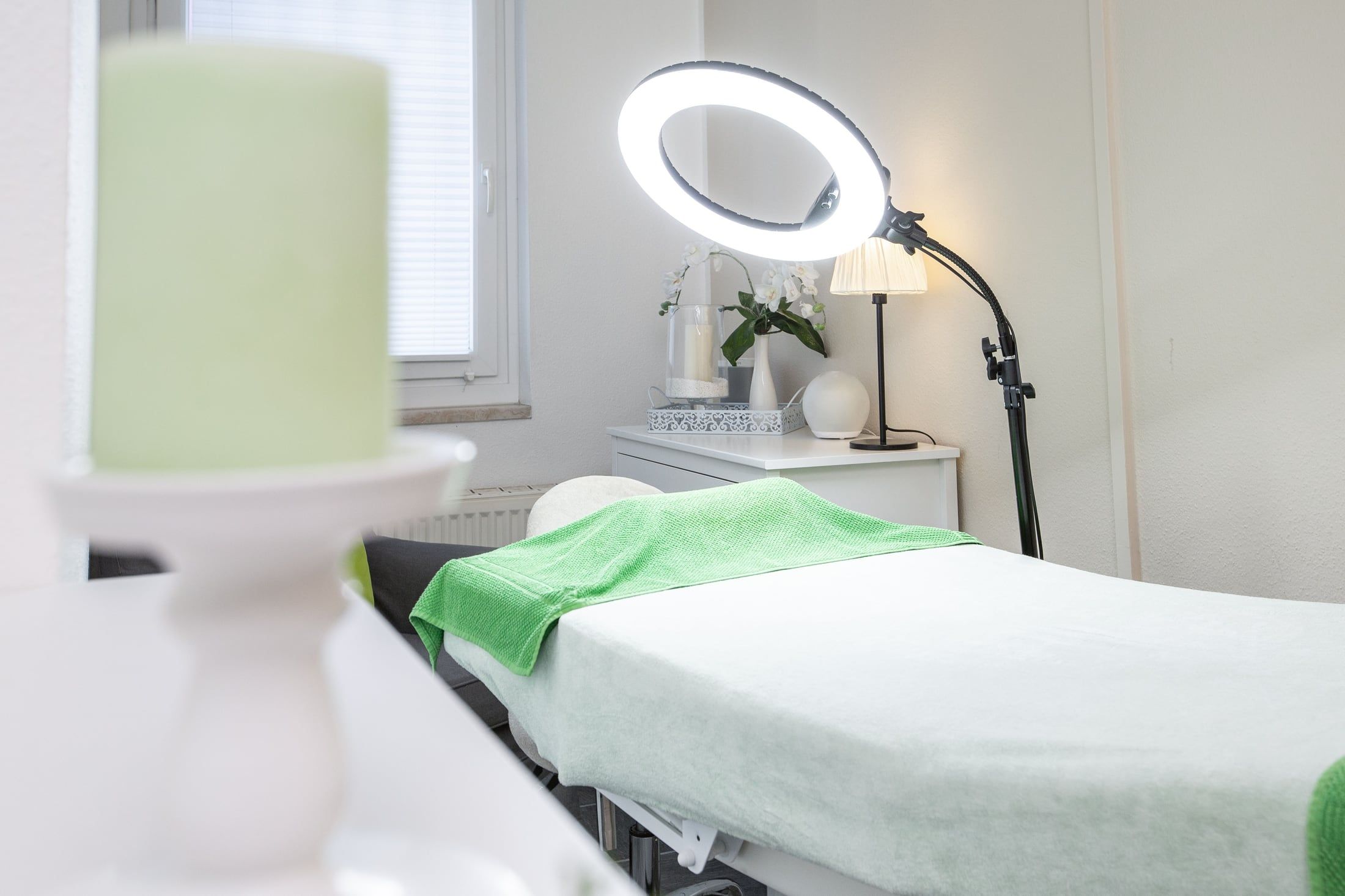 Behandlungsraum mit Kosmetikliege, grünem Handtuch, Ringleuchte, Orchidee und Kerze im Vordergrund.