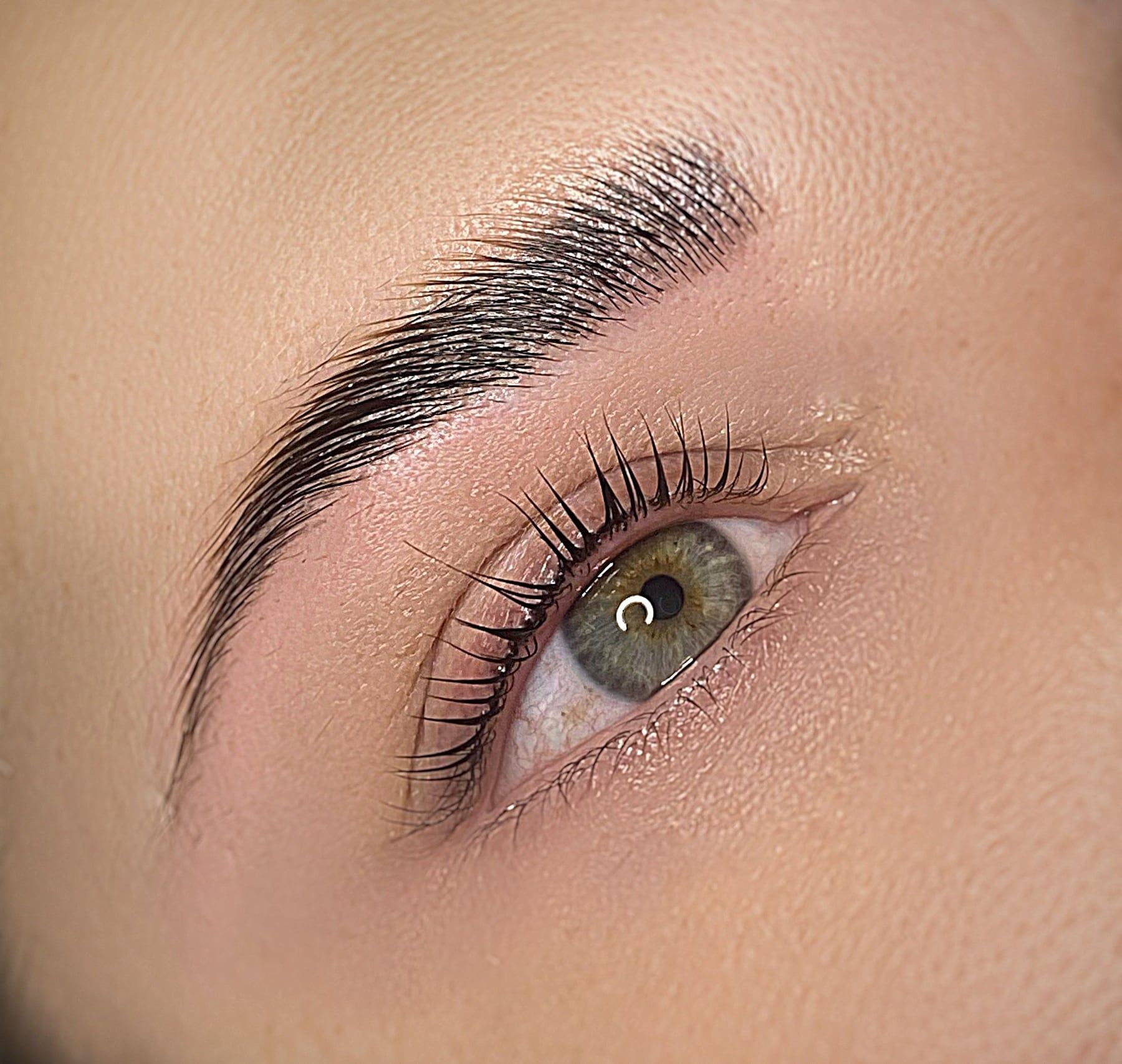 Nahaufnahme eines geöffneten grünen Auges mit dichter, nach oben gebürsteter Augenbraue und geschwungenen Wimpern.