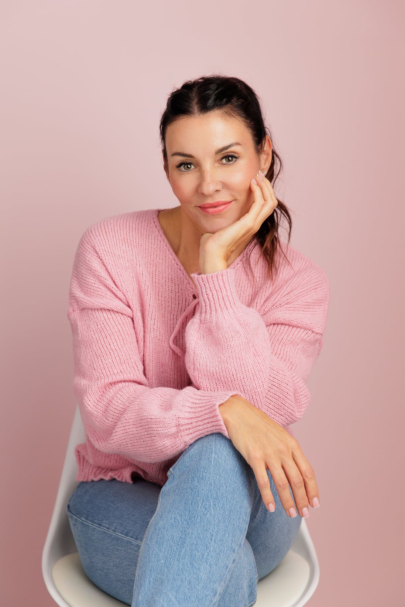 Sabrina Lindenau im rosa Strickpullover auf einem weißen Stuhl vor rosa Hintergrund, den Kopf auf die Hand gestützt.